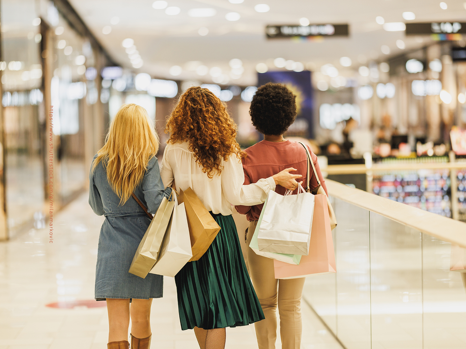Three women walk down a walkway inside a shopping mall side-by-side.